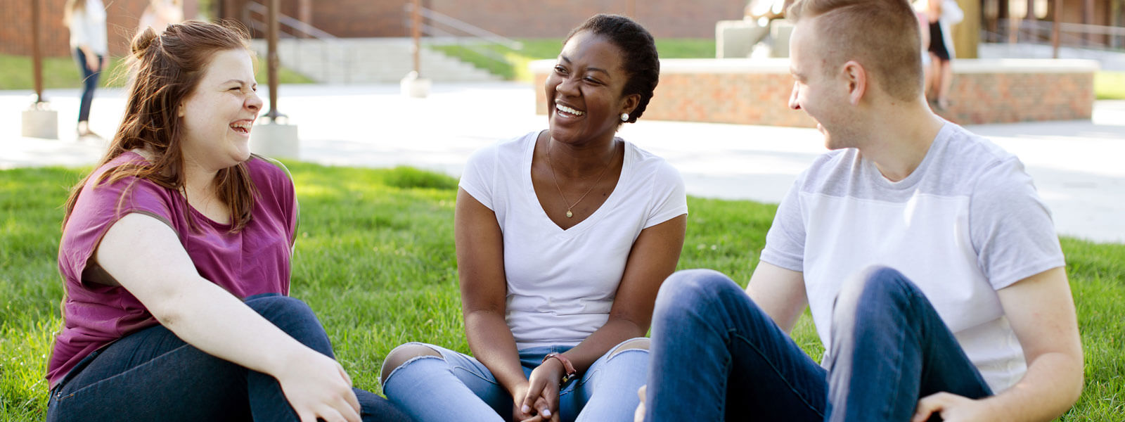 Two female students and one male student sitting on grass on forum laughing